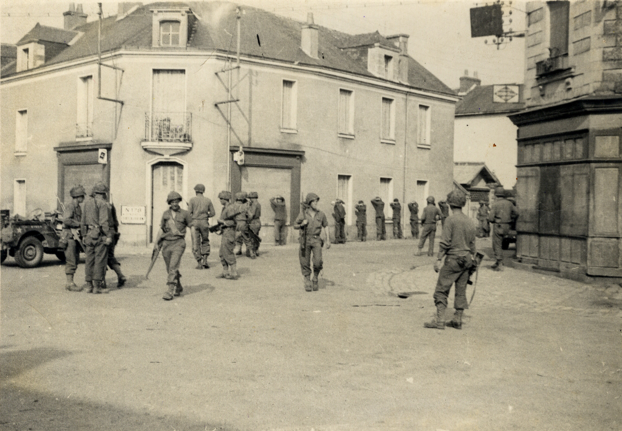 Arrivée des Américains à Carquefou le 9 août 1944. Au premier plan des soldats américains arme à la main, une jeep. A l'arrière plan des prisonniers alignés le long d'un mur (26Fi1231)