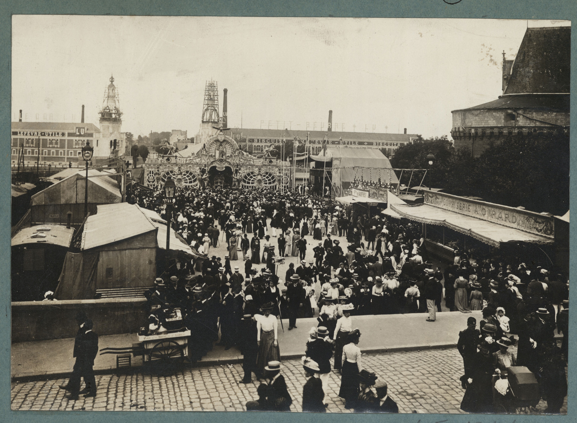 La foire d’été de 1908 sur la place de la Duchesse Anne. À l'arrière-plan, vue sur les deux tours de l'usine Lefèvre-Utile en construction (26Fi1946)